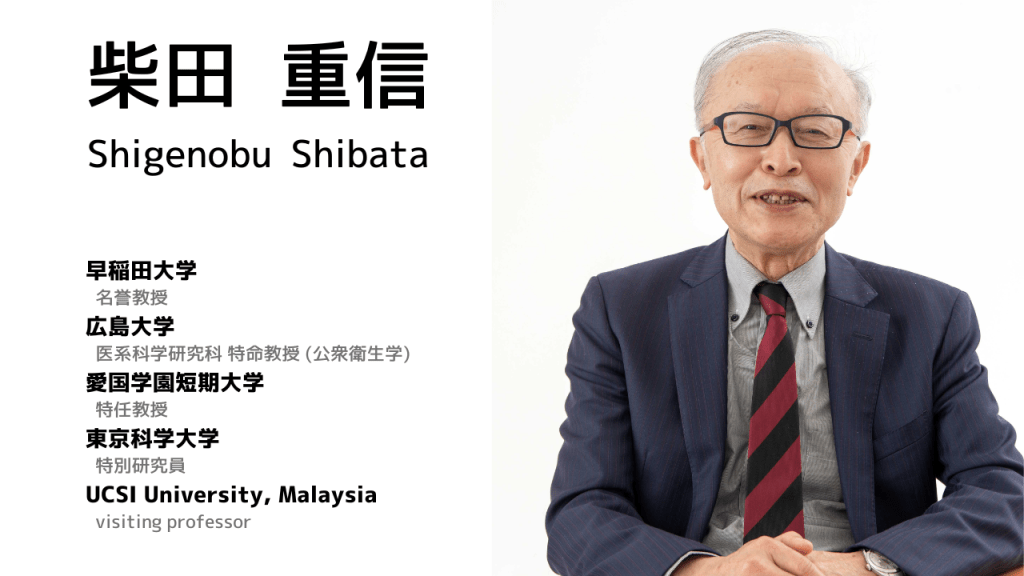 A portrait of Shigenobu Shibata, seated and smiling, wearing glasses, a black and red striped tie, and a dark blazer, in front of a plain white background.