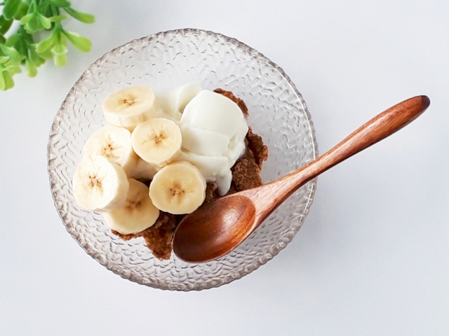 バナナとヨーグルトがトッピングされたデザートの写真。透明な皿に盛り付けられ、木製のスプーンが横に置かれている。背景には緑の植物が見える。